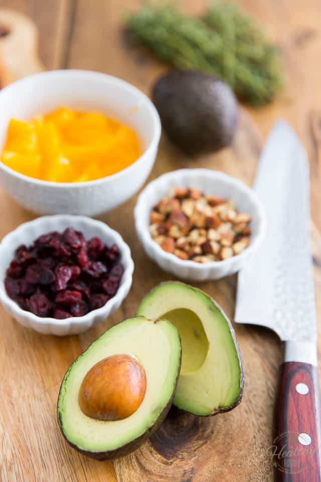 An avocado cut in half, little white bowls containing dried cranberries and toasted almonds and another one containing chunks of orange, all on a wooden cutting board with a chef knife next to them