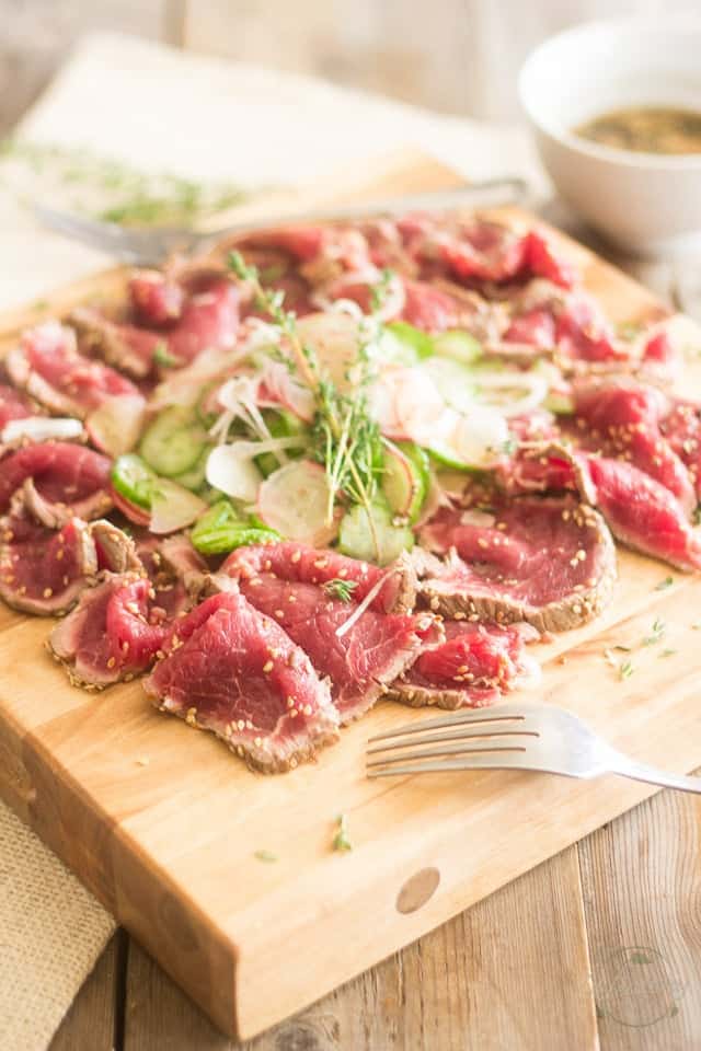 A serving of beef tataki with cucumber and radish salad served on a wooden board with a fork in the foreground