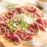 A serving of beef tataki with cucumber and radish salad served on a wooden board with a fork in the foreground