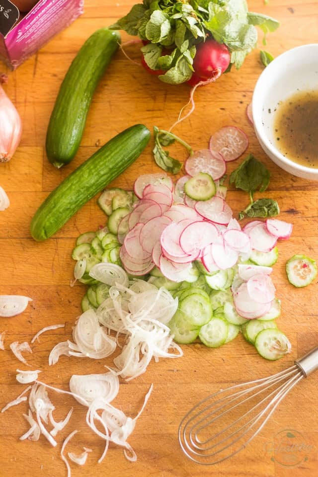 Thin slices of radish and cucumber on a wooden board, with whole cucumbers and radishes and a little bit of salad dressing in a white bowl 