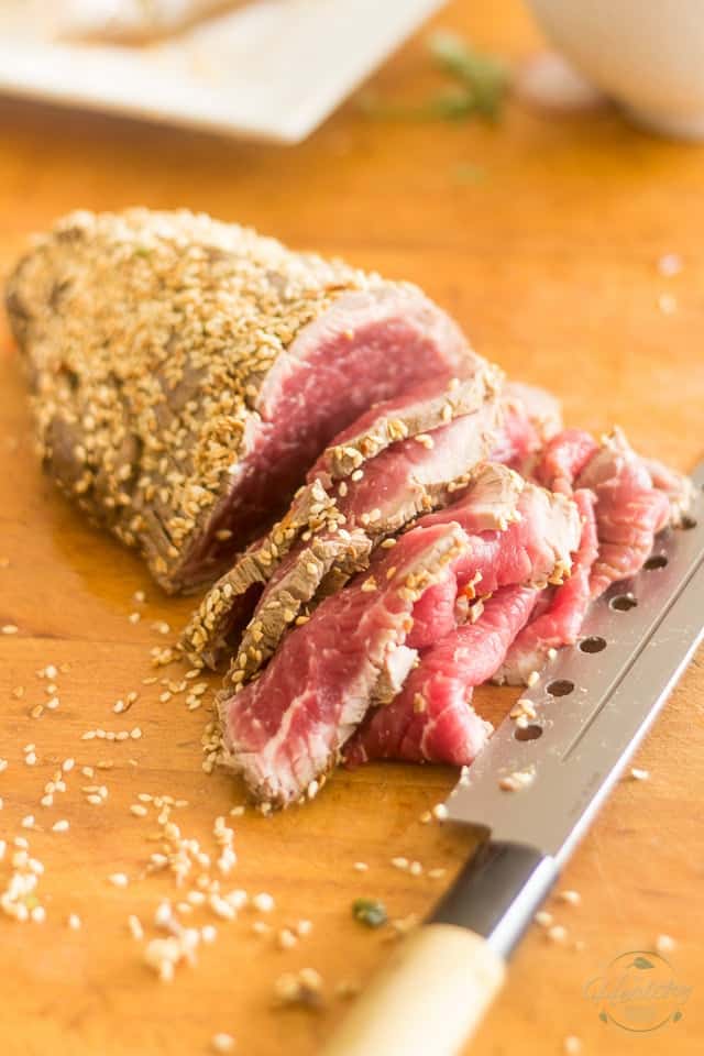 A piece of beef seared on the exterior, in the process of getting sliced on a wooden cutting board with a knife resting to its right 