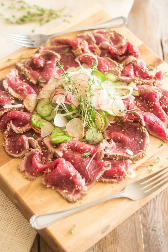 A serving of beef tataki with cucumber and radish salad served on a wooden board with a fork in the foreground