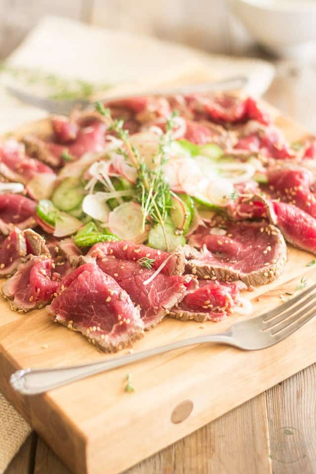 A serving of beef tataki with cucumber and radish salad served on a wooden board with a fork in the foreground