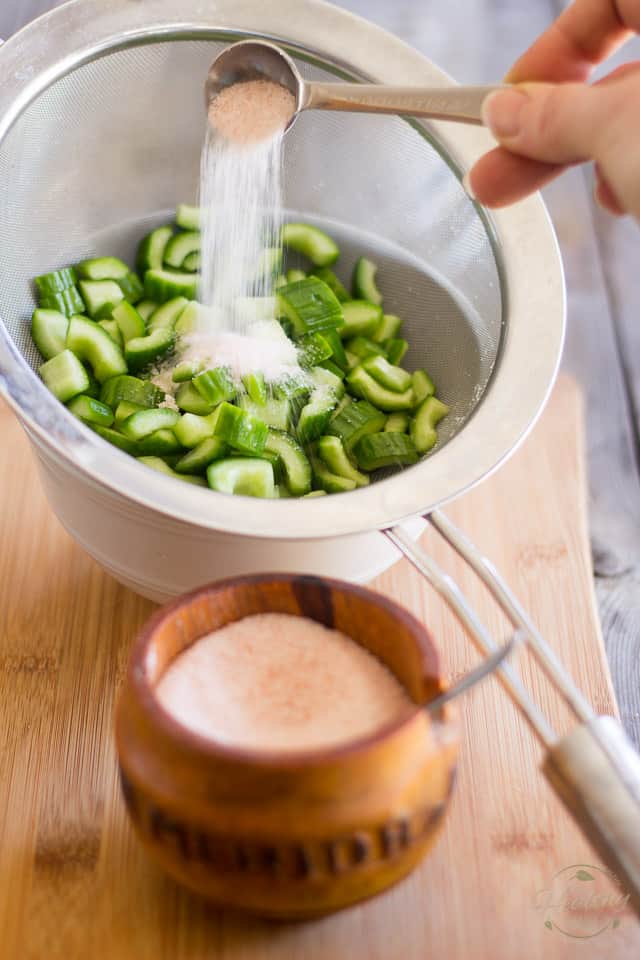 Sliced of seeded Lebanese cucumbers in a fine meshed sieve getting sprinkled with Himalayan salt