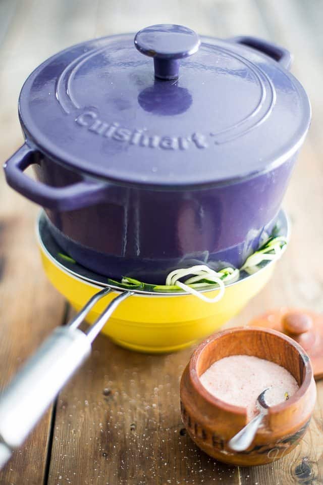 Zucchini noodles draining in a sieve set over a bowl with a purple Dutch oven on top to weigh them down
