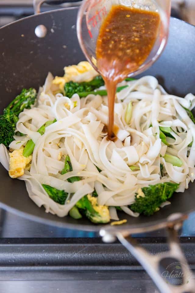 Sauce being poured into a pile of cooked rice noodles with broccolini, green onions and scrambled eggs in a black non-stick wok