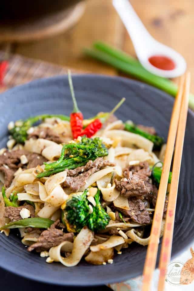 A serving of beef pad see ew in a black bowl, garnished with a couple of birds eye peppers with a pair of chopsticks resting on the rim of the bowl
