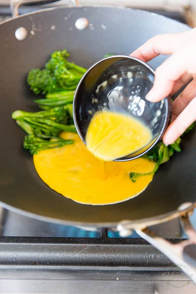 Beaten eggs in a black bowl are being poured into a non-stick wok where broccolini and green onions are already cooked and pushed to the side