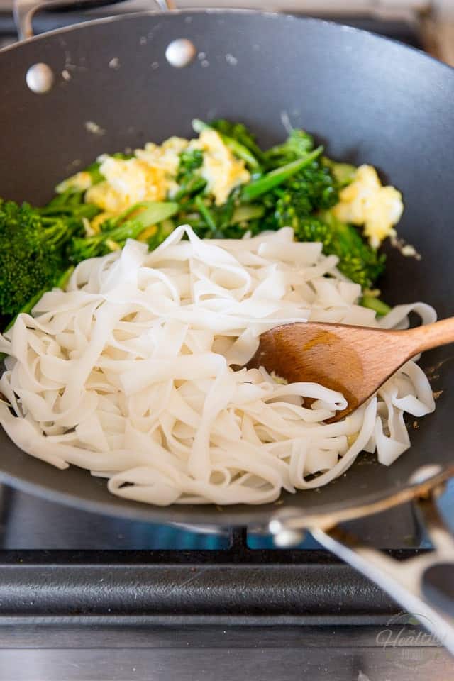 Cooked rice noodles in a non stick wok with wooden spoon and cooked broccolini and eggs in the back