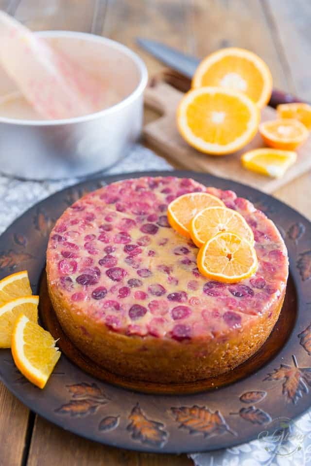 A cranberry orange cake freshly turned onto a dark brown cake plate, garnished with a few slices of orange
