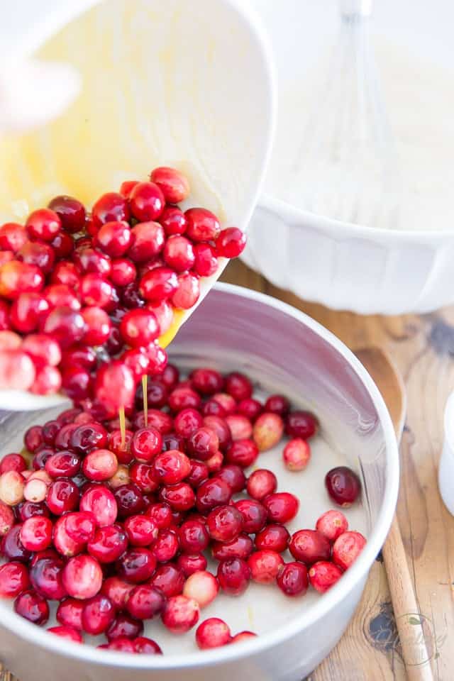 Fresh cranberries mixed with orange juice and honey being poured out of a white bowl and into an aluminum cake pan