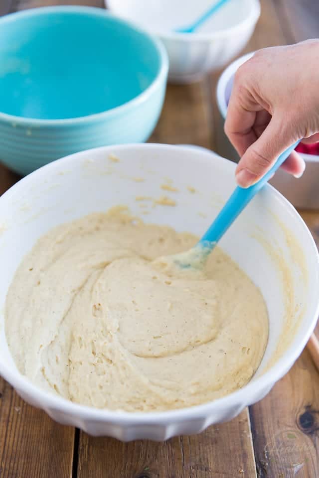 Some cake batter in a white bowl is being stirred with a blue rubber spatula