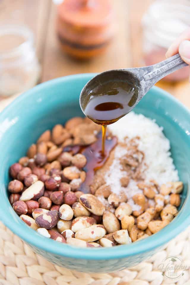 Nuts, coconut and spices in a blue bowl, over which a tablespoon of maple syrup is being poured