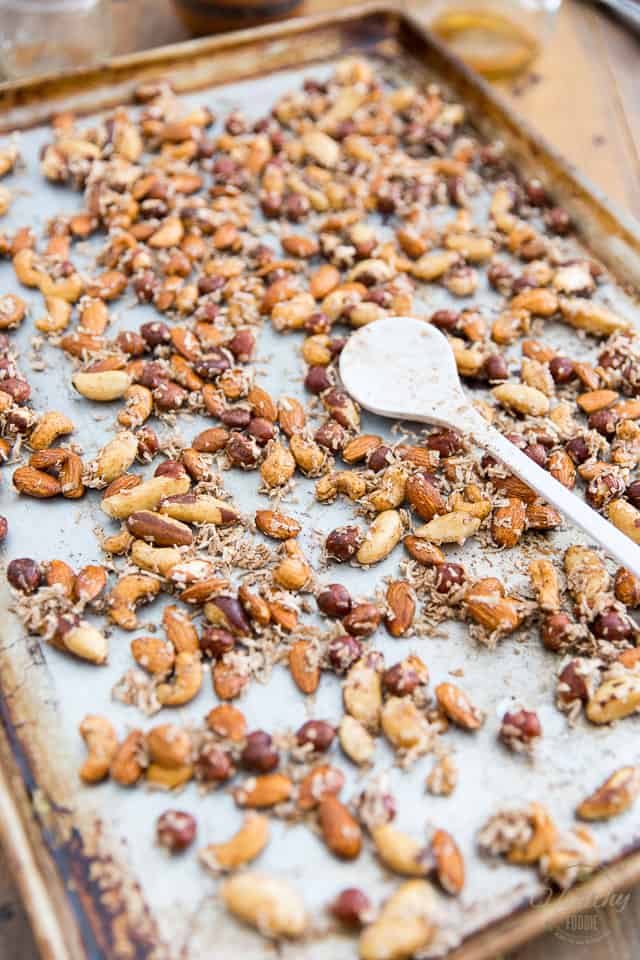 Spiced nuts and coconut on a large sheet pan with large white plastic spoon spreading nuts