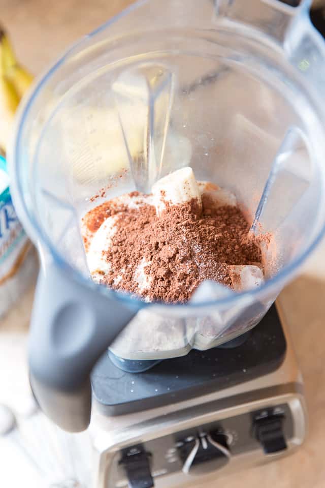 Half a banana, milk, cocoa powder, whey protein powder and ice cubes in the container of a Vitamix Speed Blender