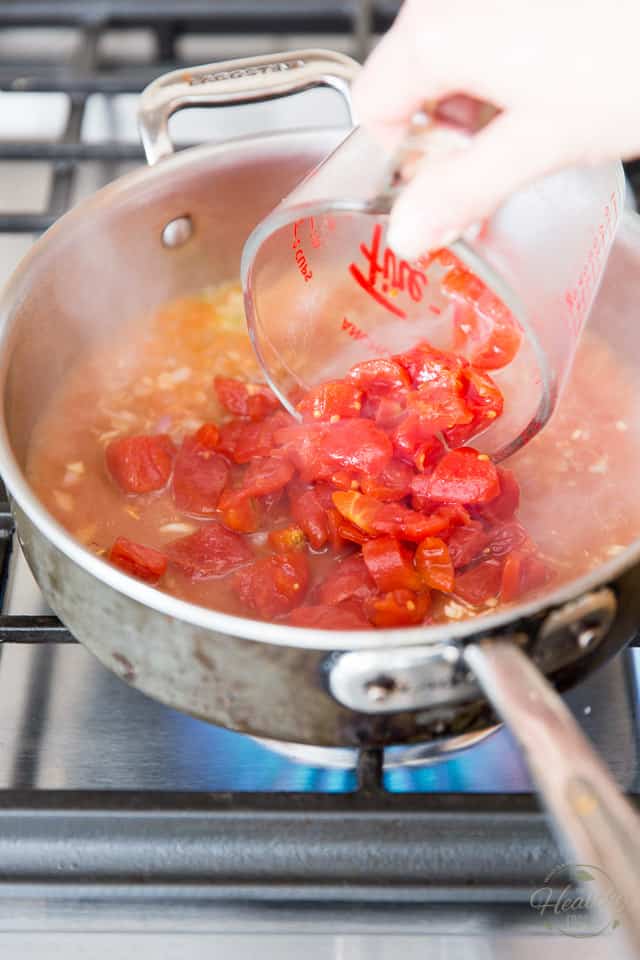 Canned diced tomatoes are being poured out of a glass measuring cup and into a stainless steel saute pan