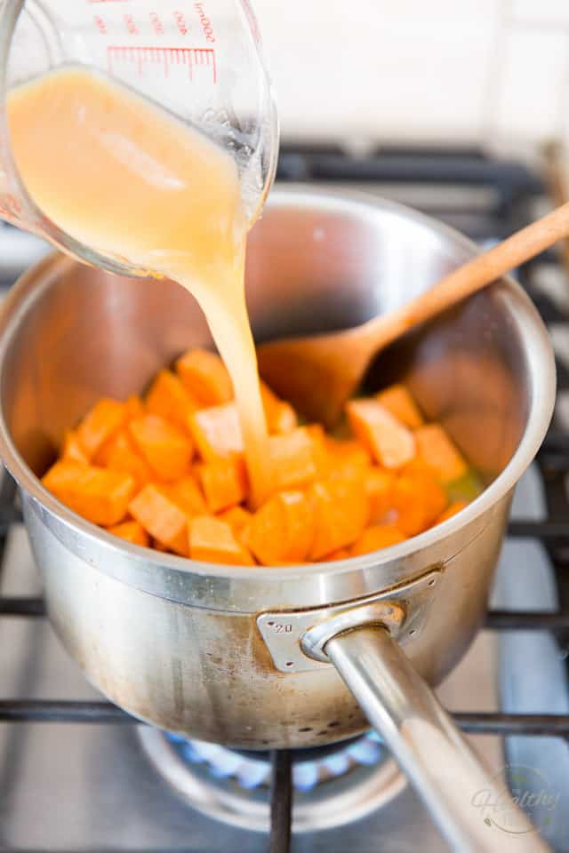 Chicken broth is being poured into a stainless steel saucepan that already contains cubes of sweet potatoes