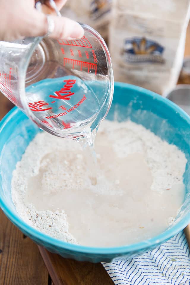 Water is being poured from a glass measuring cup into a blue bowl containing flours
