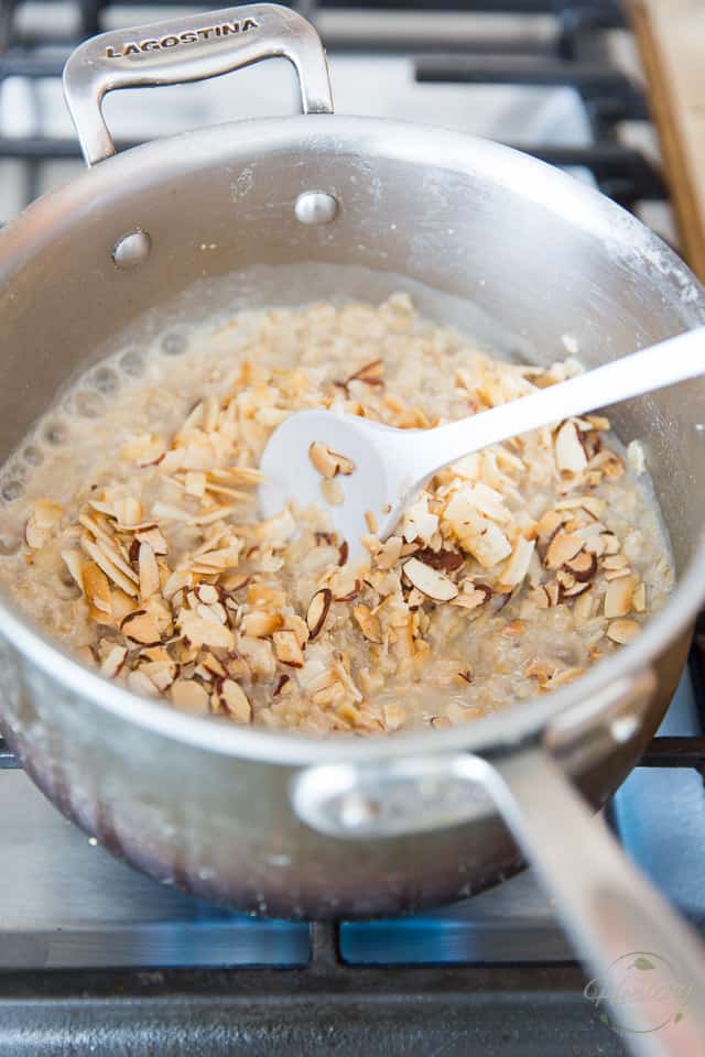 Toasted coconut and almonds are getting stirred into cooked oatmeal in a saucepan