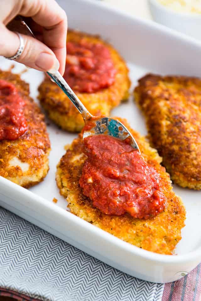 Cooked breaded chicken breasts in white baking dish getting topped with spoonfuls of marinara sauce