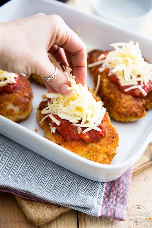 Cooked breaded chicken cutlets in a white baking dish, topped with marinara sauce, getting topped with shredded cheese