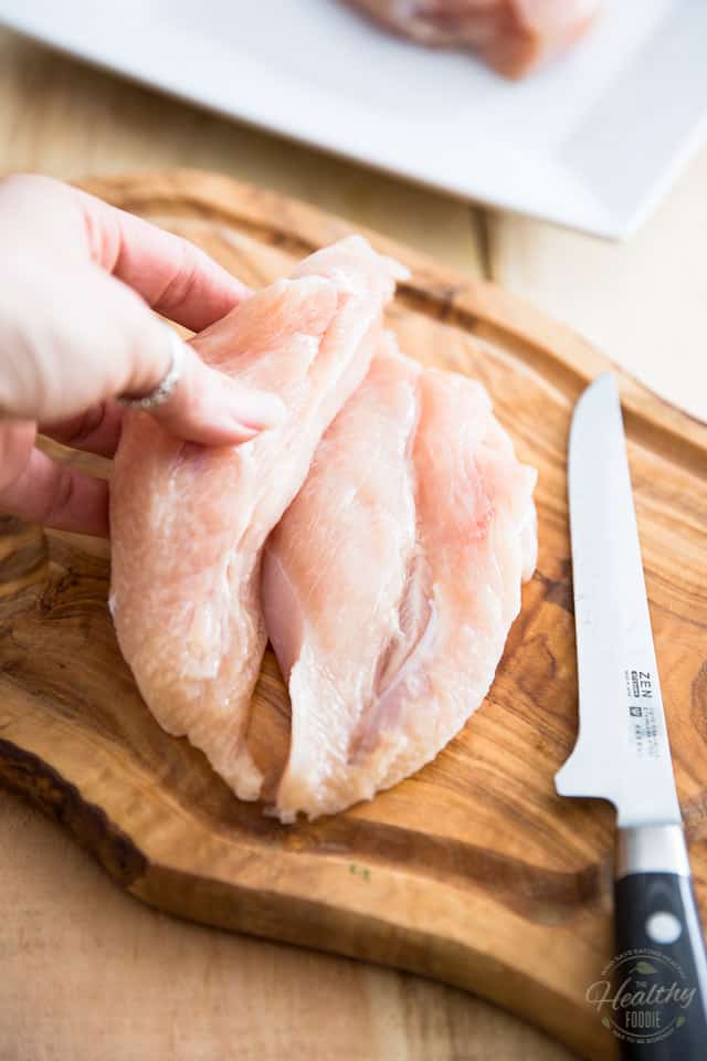 A chicken breast sliced in half lengthwise on a wooden cutting board with boning knife next to it