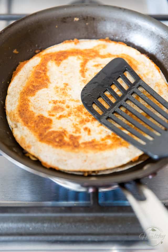 A chicken quesadilla cooking in a non-stick skillet getting pressed down with a black spatula