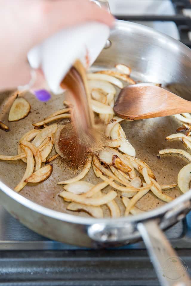 A blend of spices is being poured over sauteed onions in a stainless steel skillet