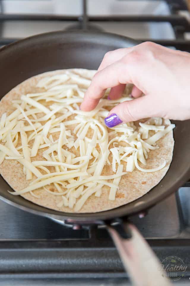 Some shredded cheese is getting dispersed over a flour tortilla placed in a non-stick pan over gas burner