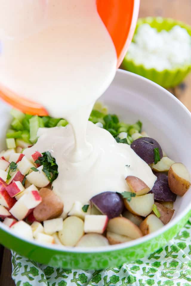 A creamy dressing is being poured out of an orange bowl and into a bowl containing cooked potatoes, apples, celery, green onions and parsley