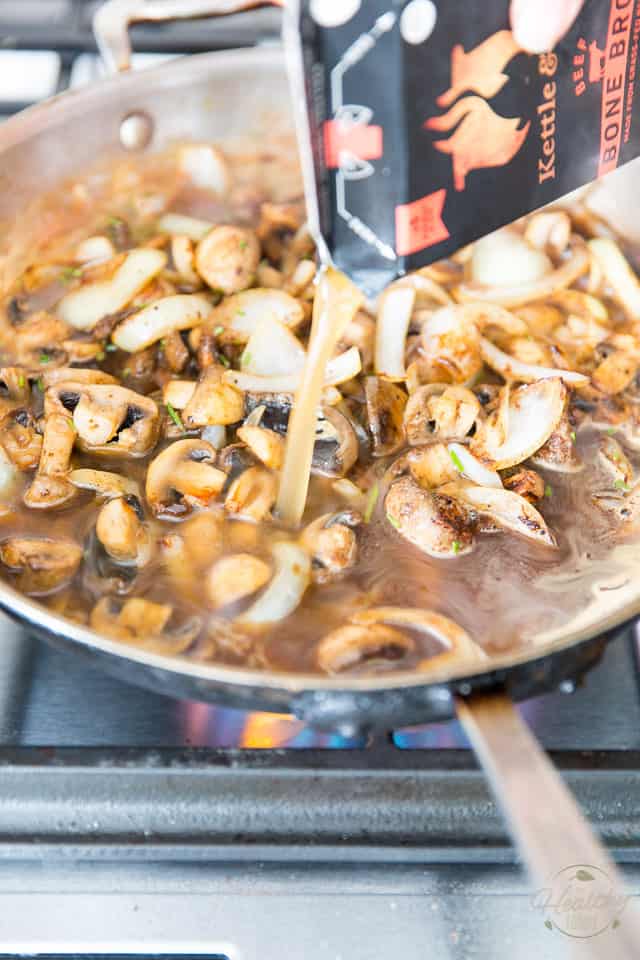 Bone broth is being poured into a stainless steel skillet containing sauteed onions and mushrooms