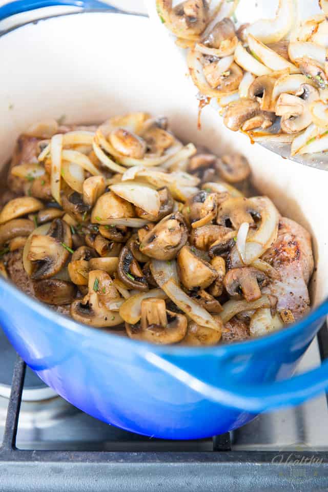 Sauteed onions and mushrooms are being added to a blue Dutch oven already containing braised lamb meat