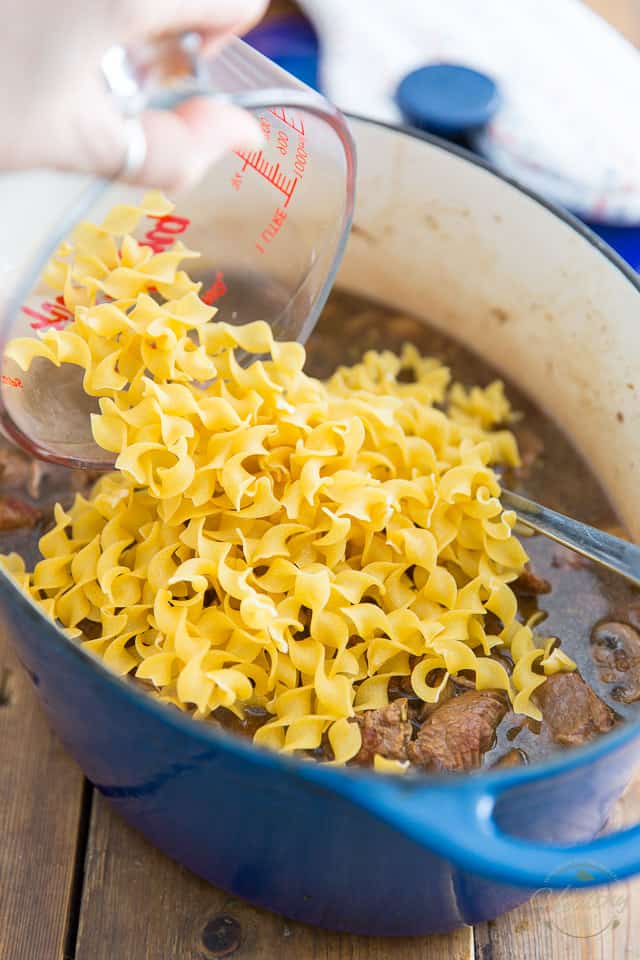 Adding dry egg noodles to a blue Dutch oven containing braised meat and cooking liquid