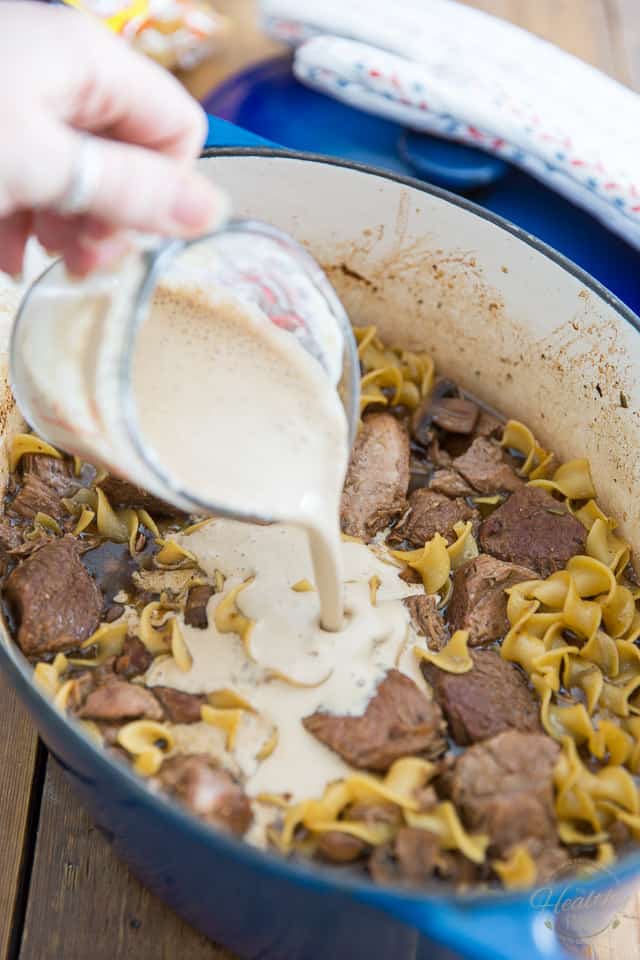 A mixture of sour cream, beer and corn starch is being poured into a blue Dutch oven containing some Lamb and Mushroom Ragu