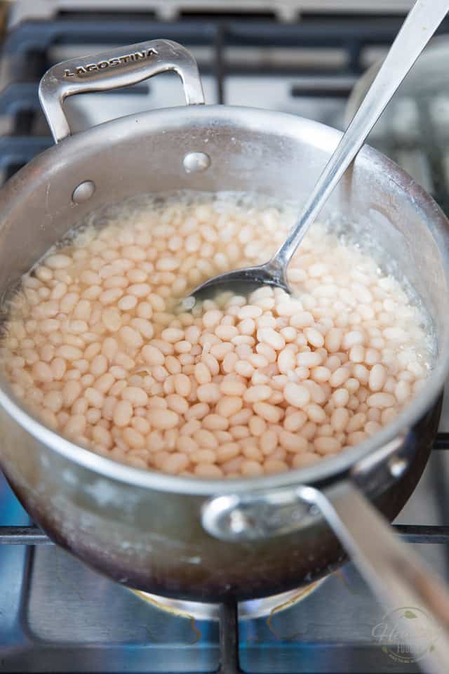 White navy beans cooking on a gas range in a medium saucepan with large stainless steel spoon sticking out