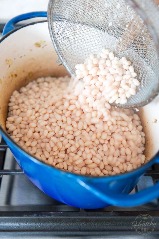 Cooked white beans are being poured from a fine meshed strainer and into a blue Dutch oven