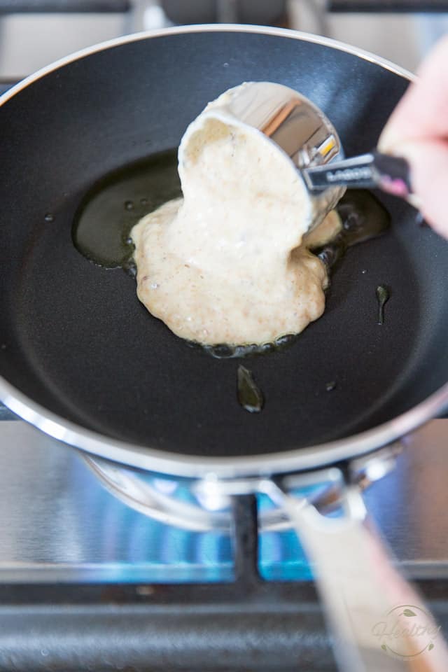 Pancake batter is getting poured out of a stainless steel measuring cup and into a small non-stick skillet