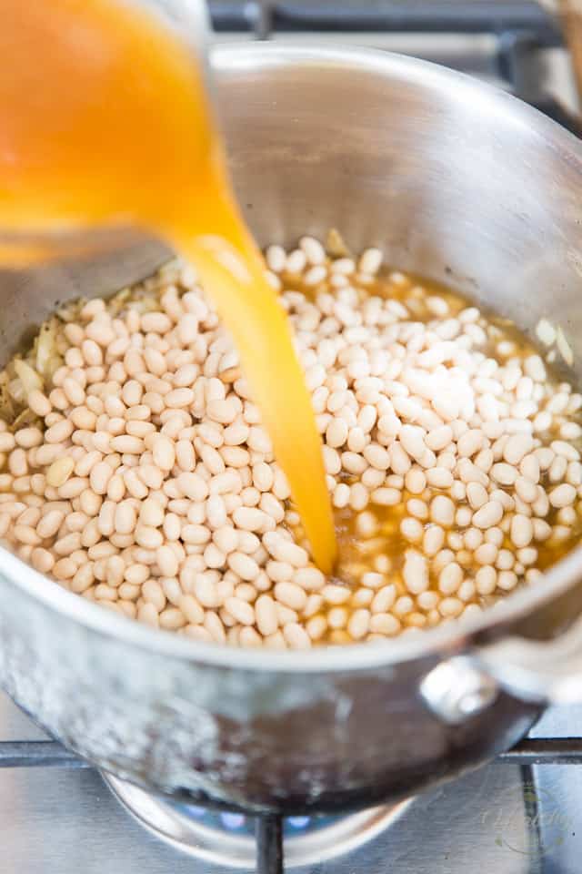 Stock is getting poured out of a glass measuring cup and into a large stock pot containing uncooked navy beans