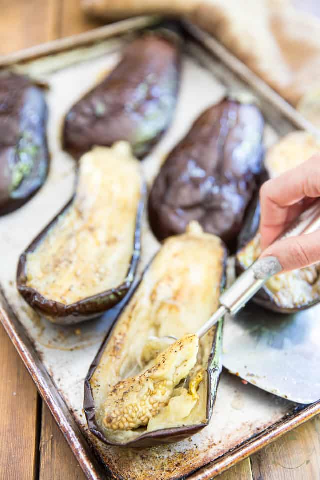 Cooked eggplants sliced in half lengthwise arranged in a baking sheet, flesh being scooped out of one of them with an ice cream spoon