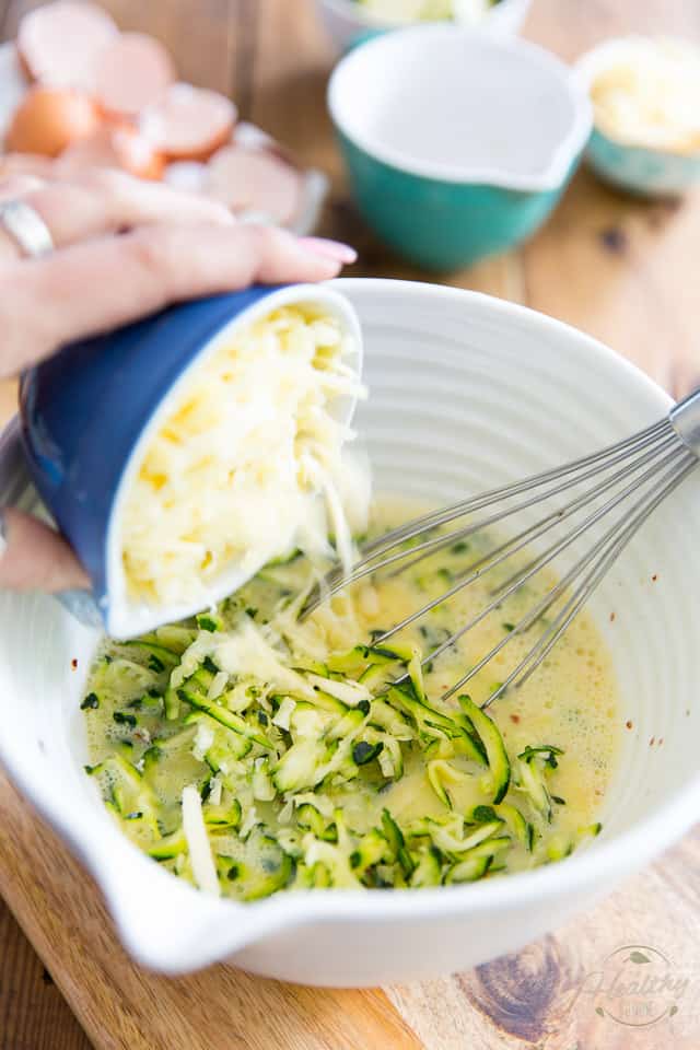 Grated Cheese getting added to white bowl containing grated zucchini and beaten eggs