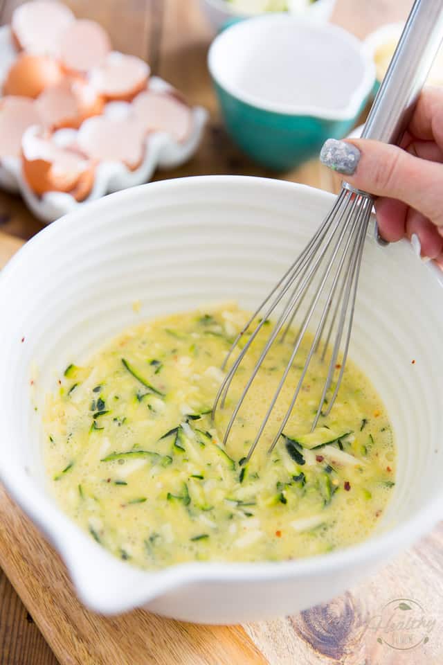 Eggs, grated cheese and grated zucchini in large white bowl getting mixed with a whisk