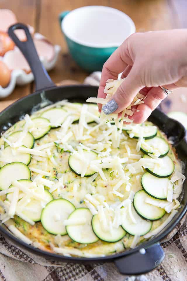 Grated cheese and sliced zucchini getting arranged on top of partially cooked zucchini frittata in cast iron skillet