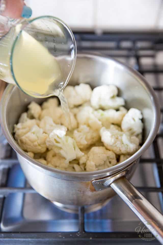 Chicken broth is being poured into a stainless steel saucepan that contains chopped cauliflower florets