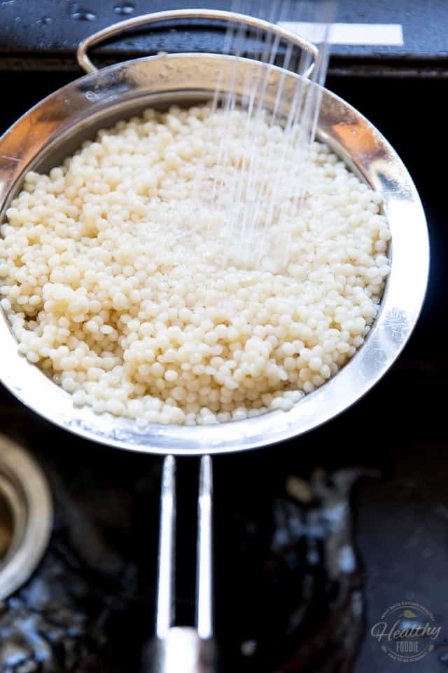 Cooked pearl couscous getting rinsed in a sieve set over a black sink