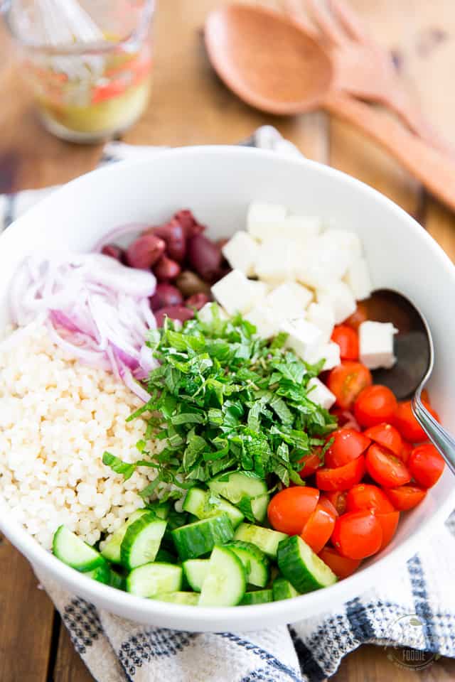 Chopped tomatoes, cucumbers, sliced red onions, kalamata olives, cubed feta cheese, cooked pearl couscous and fresh herbs in a white salad bowl