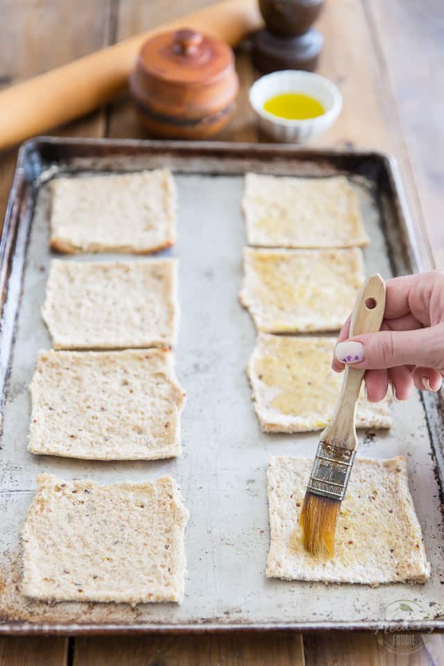Flattened pieces of bread arranged on a baking sheet are getting brushed with a little bit of olive oil