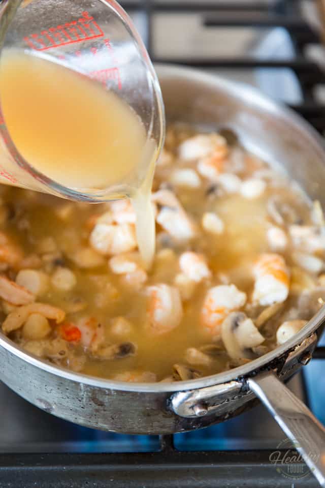 Chicken broth is being poured into a pan containing mushrooms shrimps and scallops