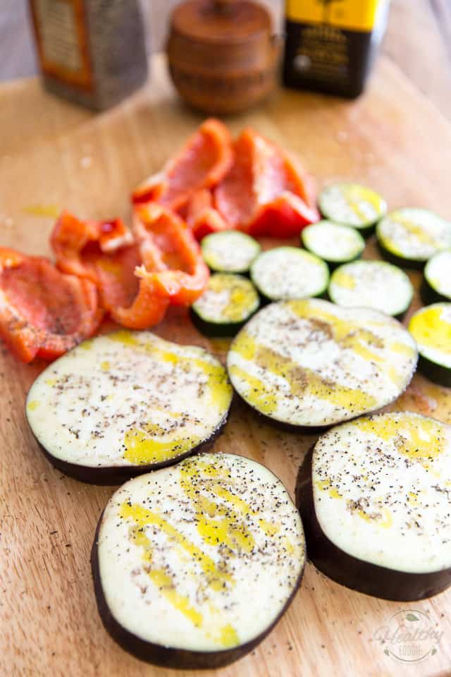 Slices of eggplant and zucchini and a red bell pepper seasoned with olive oil, salt and pepper on a wooden cutting board