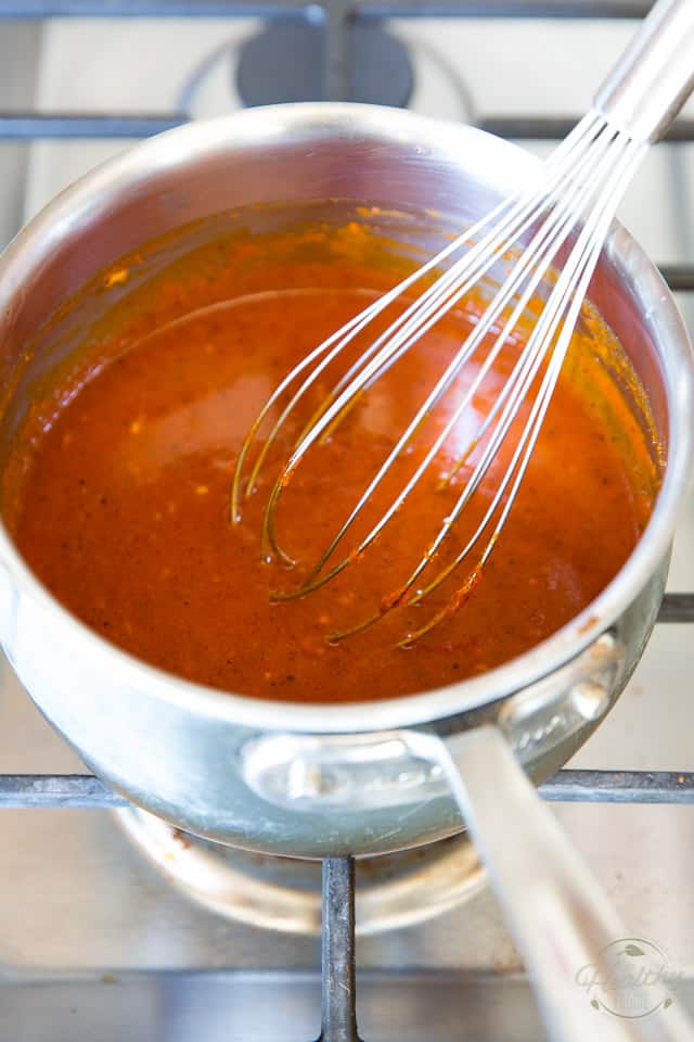 Buffalo sauce simmering on a gas stove in a small saucepan with whisk sticking out 