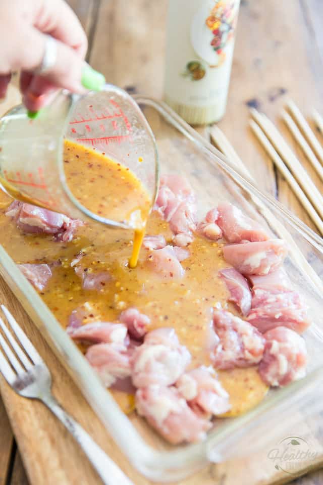 A honey dijon marinade is being poured out of a glass measuring cup over pieces of raw chicken in a glass baking pan